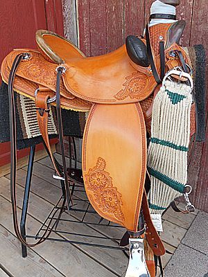 Tan leather western saddle on a metal stand, with tooling, against a red wooden backdrop.