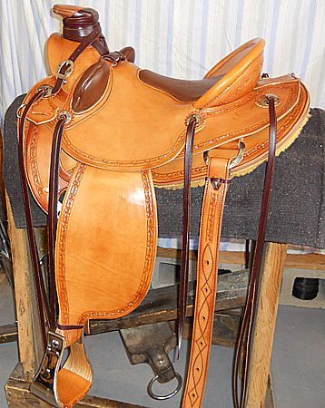 Tan leather western saddle on a wooden stand, with tooled detailing, brown seat, and stirrups.