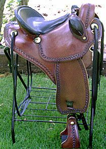 Brown leather western saddle on a black metal stand, outdoors on grass.