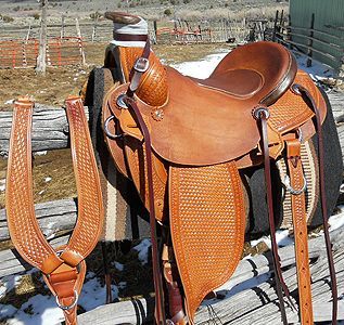 Brown leather western saddle with breast collar, resting on a wooden fence in a snowy outdoor setting.