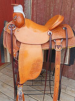 Tan leather western saddle with floral tooling, displayed on a metal stand.