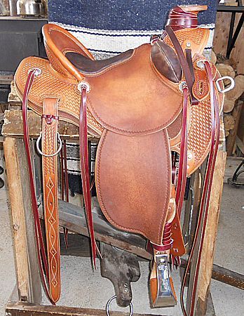 Brown leather western saddle on a wooden stand, intricate tooling details, red accents.