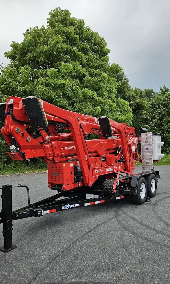 Red tree-trimming machine on a trailer, parked on asphalt. Green foliage in background.