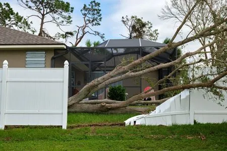A fallen tree rests on a white vinyl fence and a screened-in back porch after a storm.