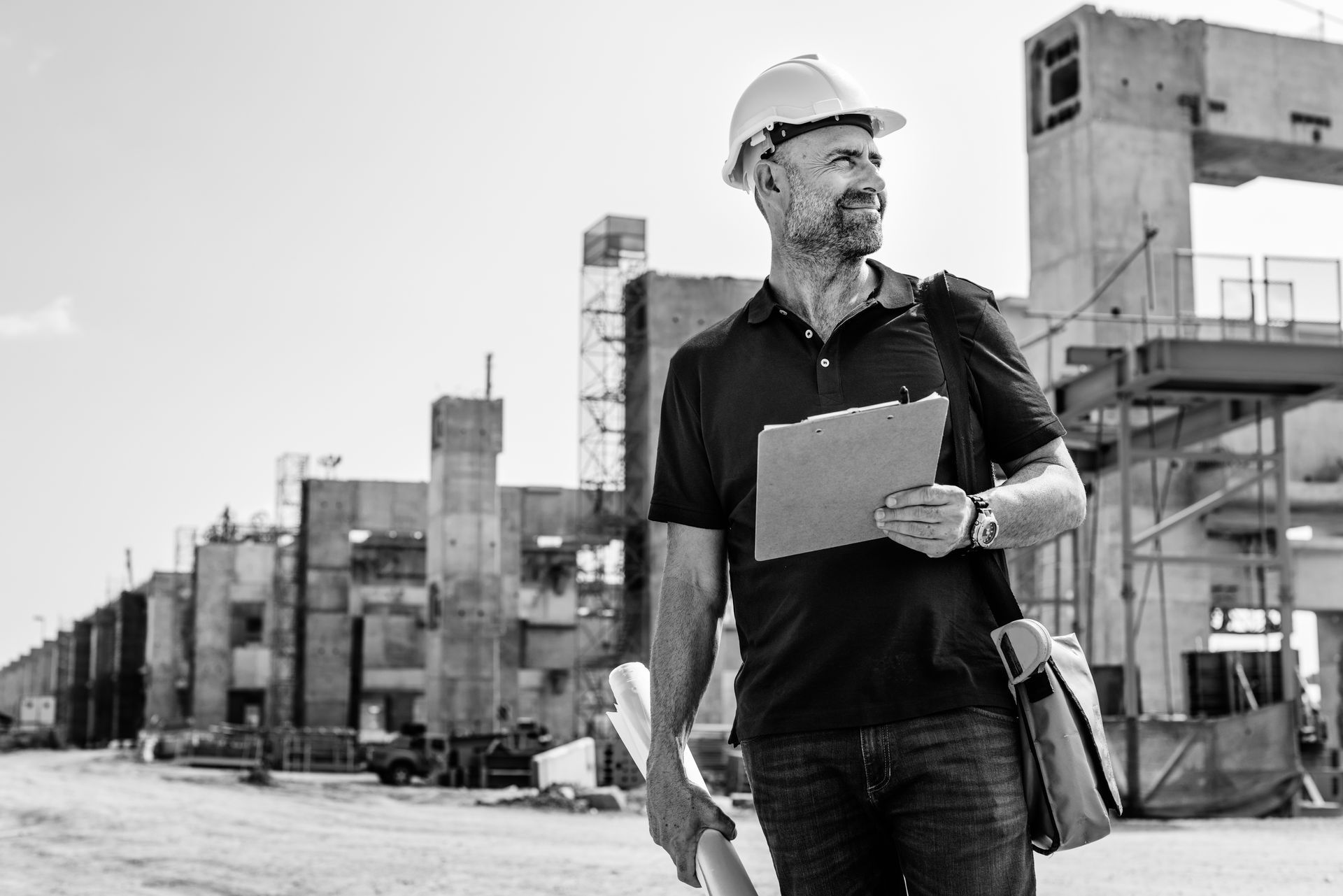 A man wearing a hard hat and holding a clipboard is walking on a construction site.