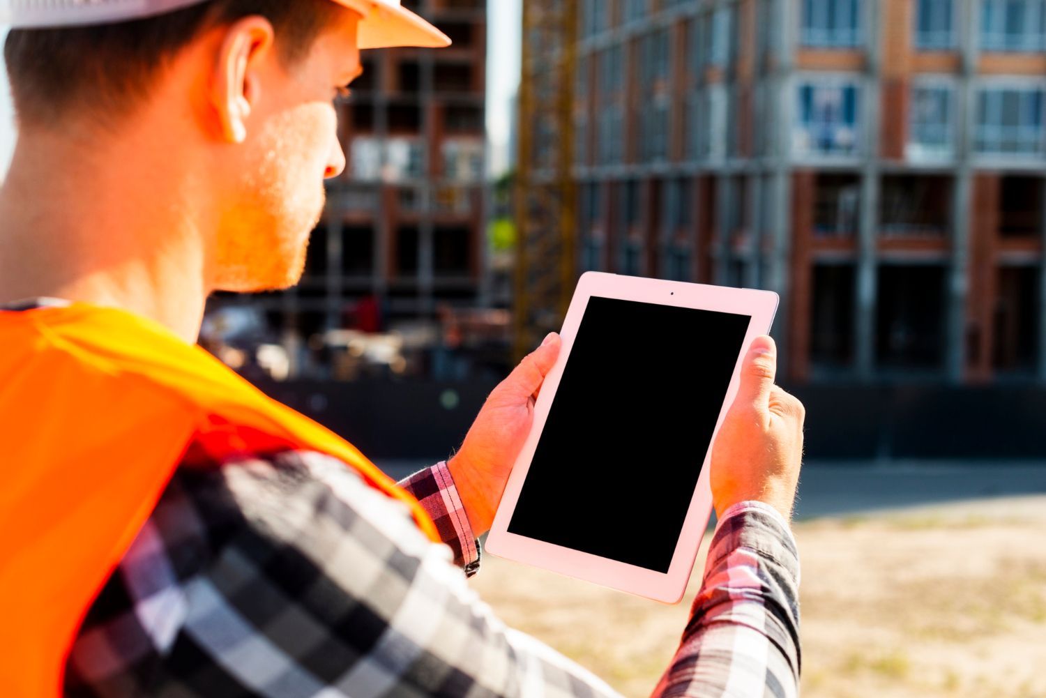 A construction worker is holding a tablet in front of a building under construction.