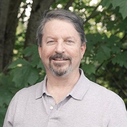 A man with a beard is smiling for the camera while standing in front of trees.