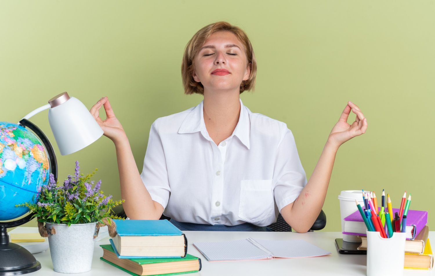 A woman is sitting at a desk with her eyes closed and meditating.