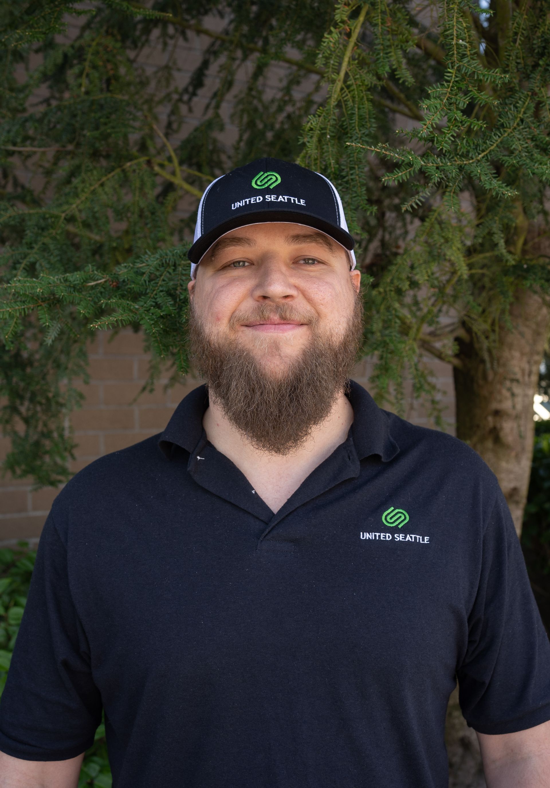 A man with a beard wearing a hat and a black shirt is standing in front of a tree.