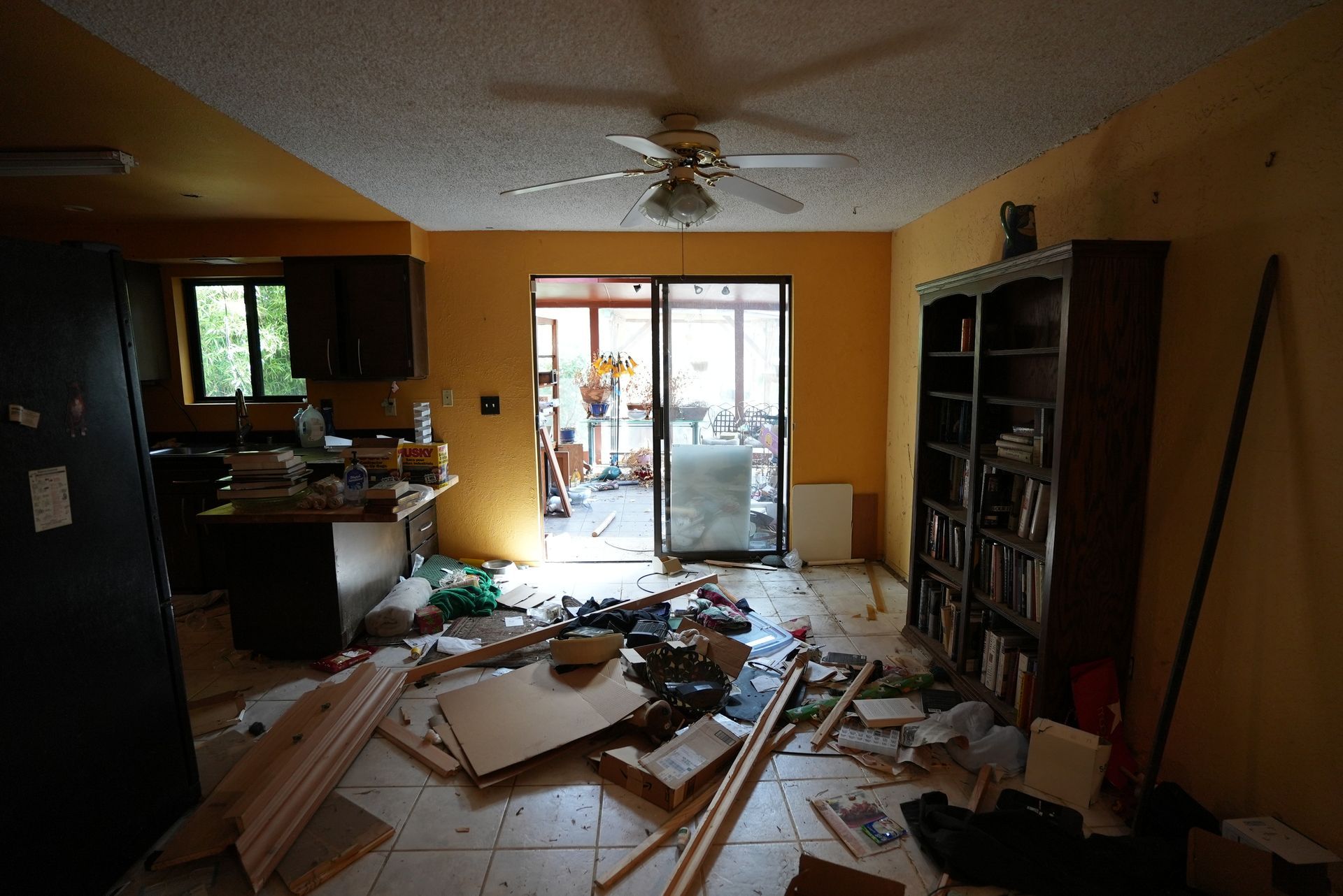 A messy living room with a ceiling fan and bookshelves