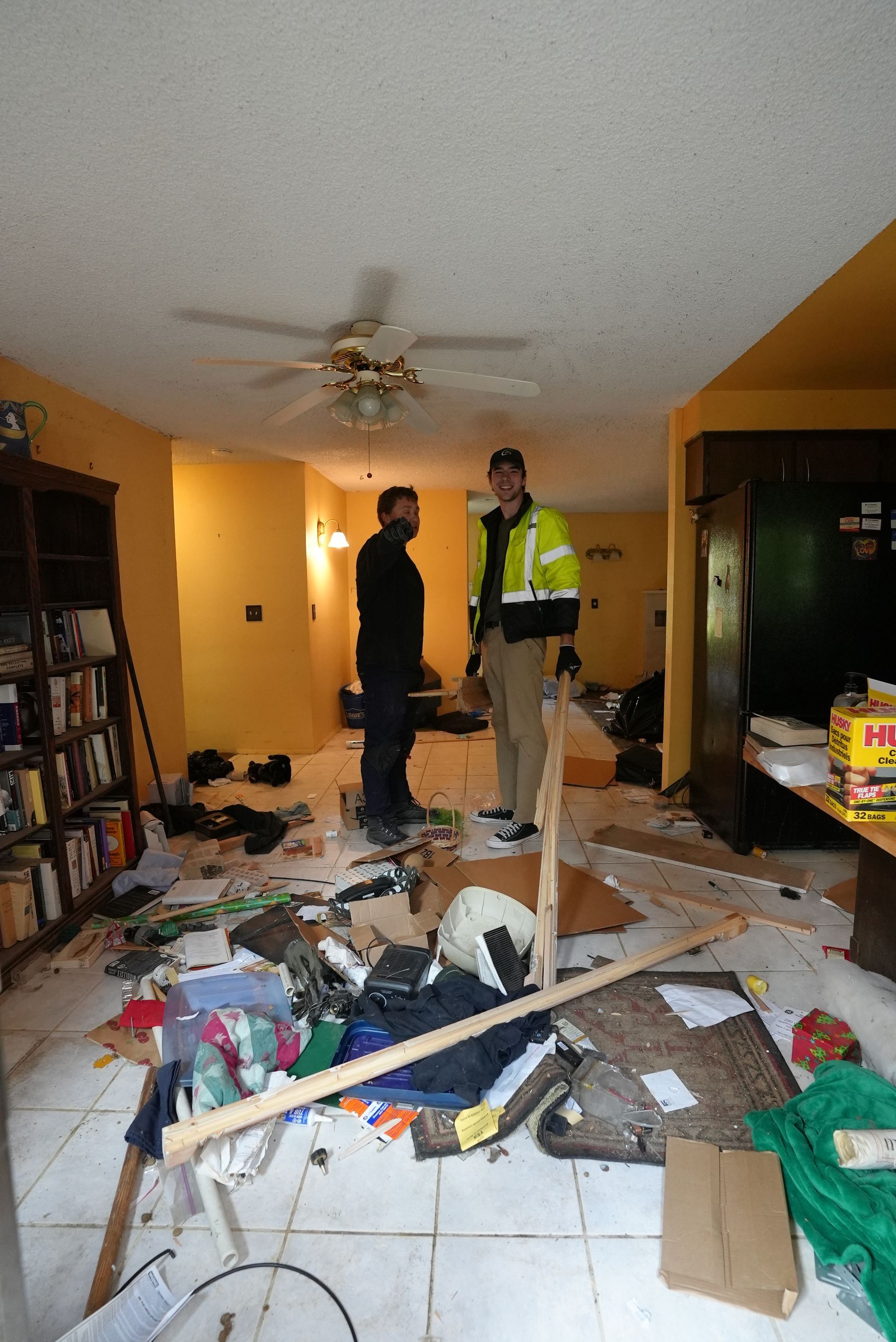 Two men are standing in a messy living room.
