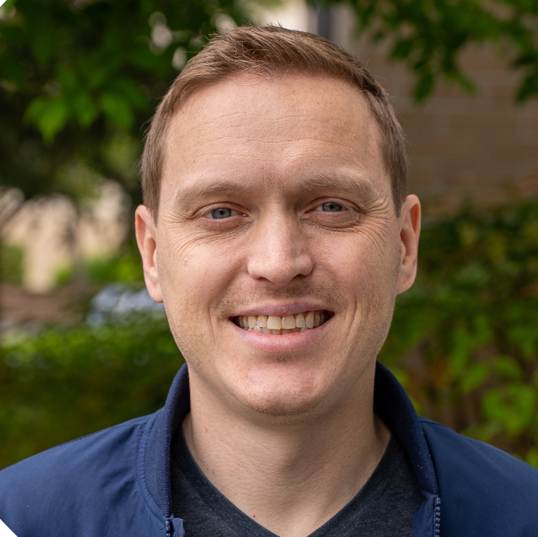 A man in a grey sweater is smiling in front of a bush.