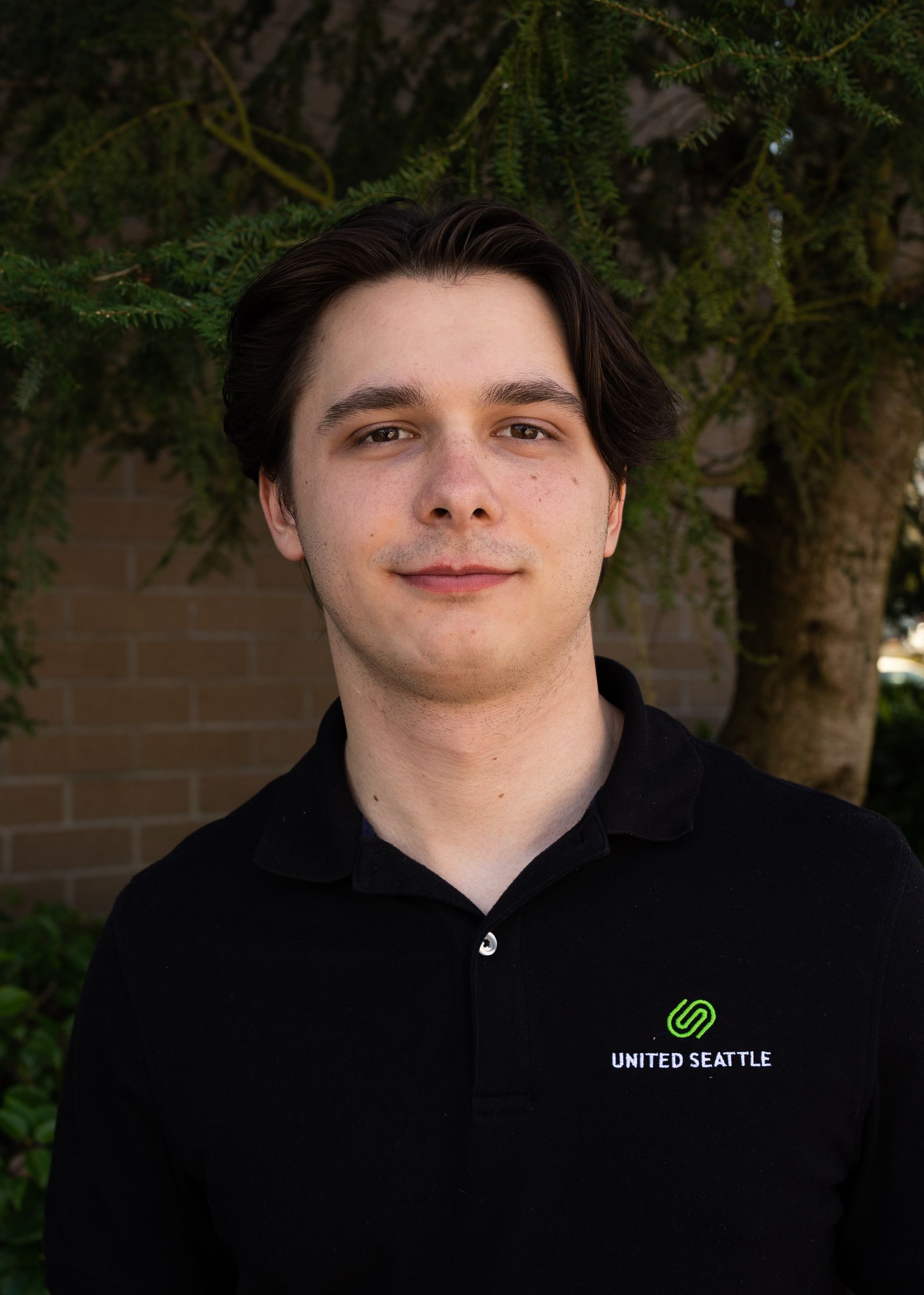 A young man in a black polo shirt is standing in front of a tree.