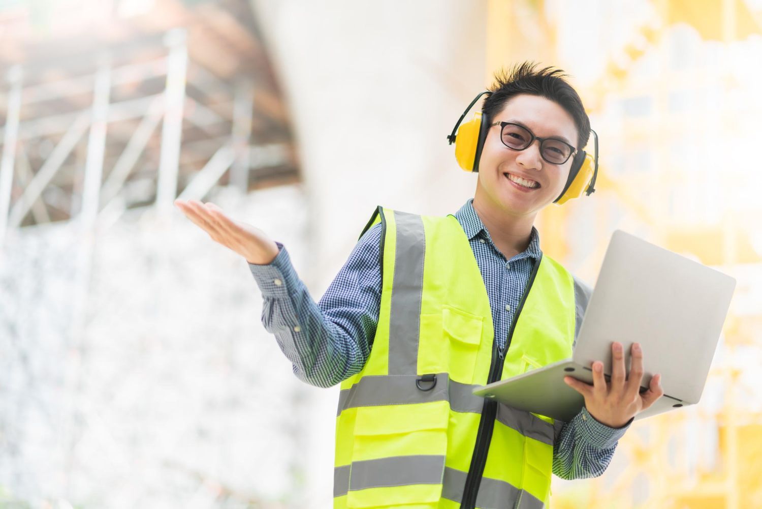 A man wearing headphones and a yellow vest is holding a laptop computer.