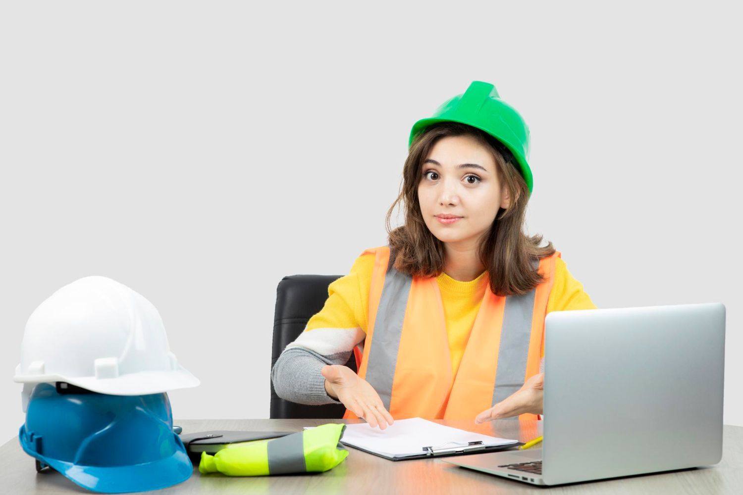 A woman is sitting at a desk with a laptop and hard hats.