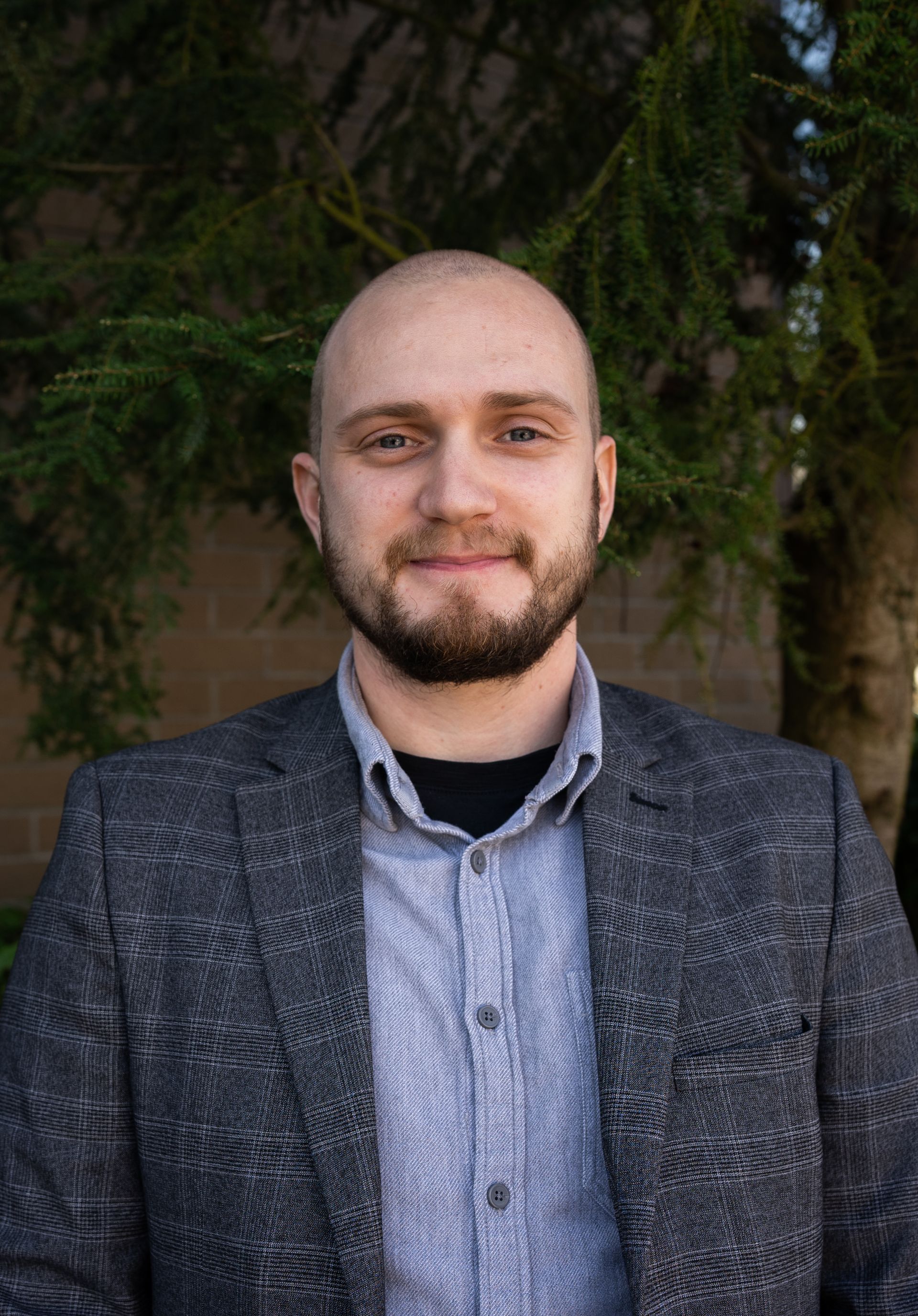 A bald man with a beard is wearing a suit and standing in front of a tree.