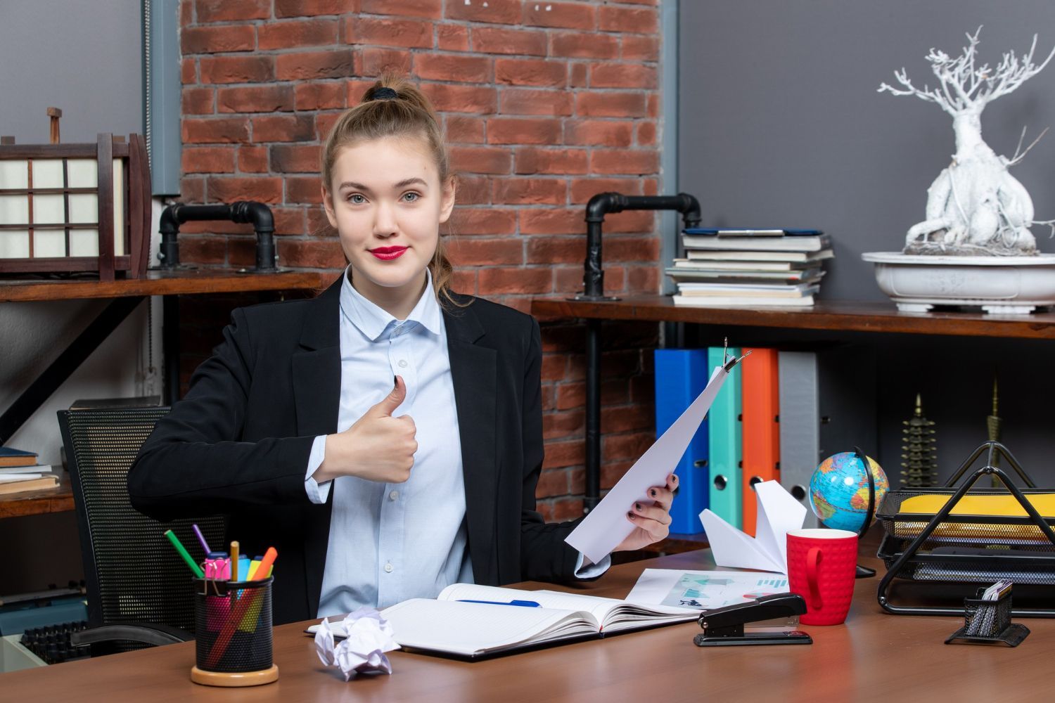 A woman is sitting at a desk in an office holding a piece of paper and giving a thumbs up.