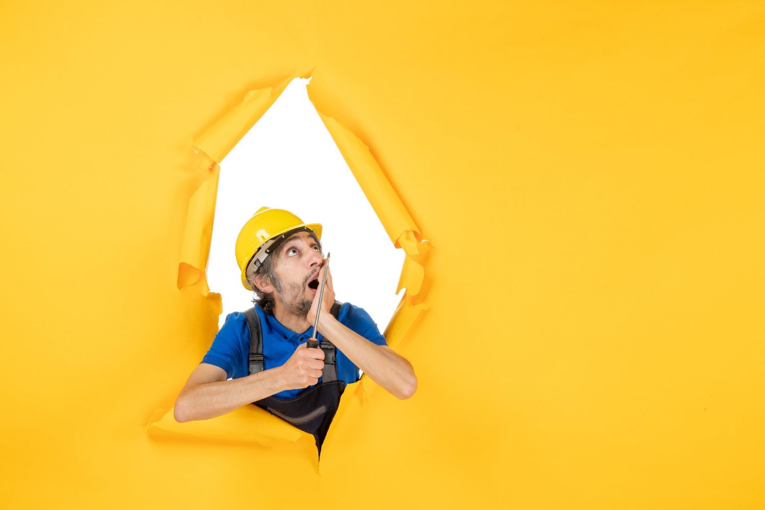 A construction worker is looking through a hole in a yellow wall.