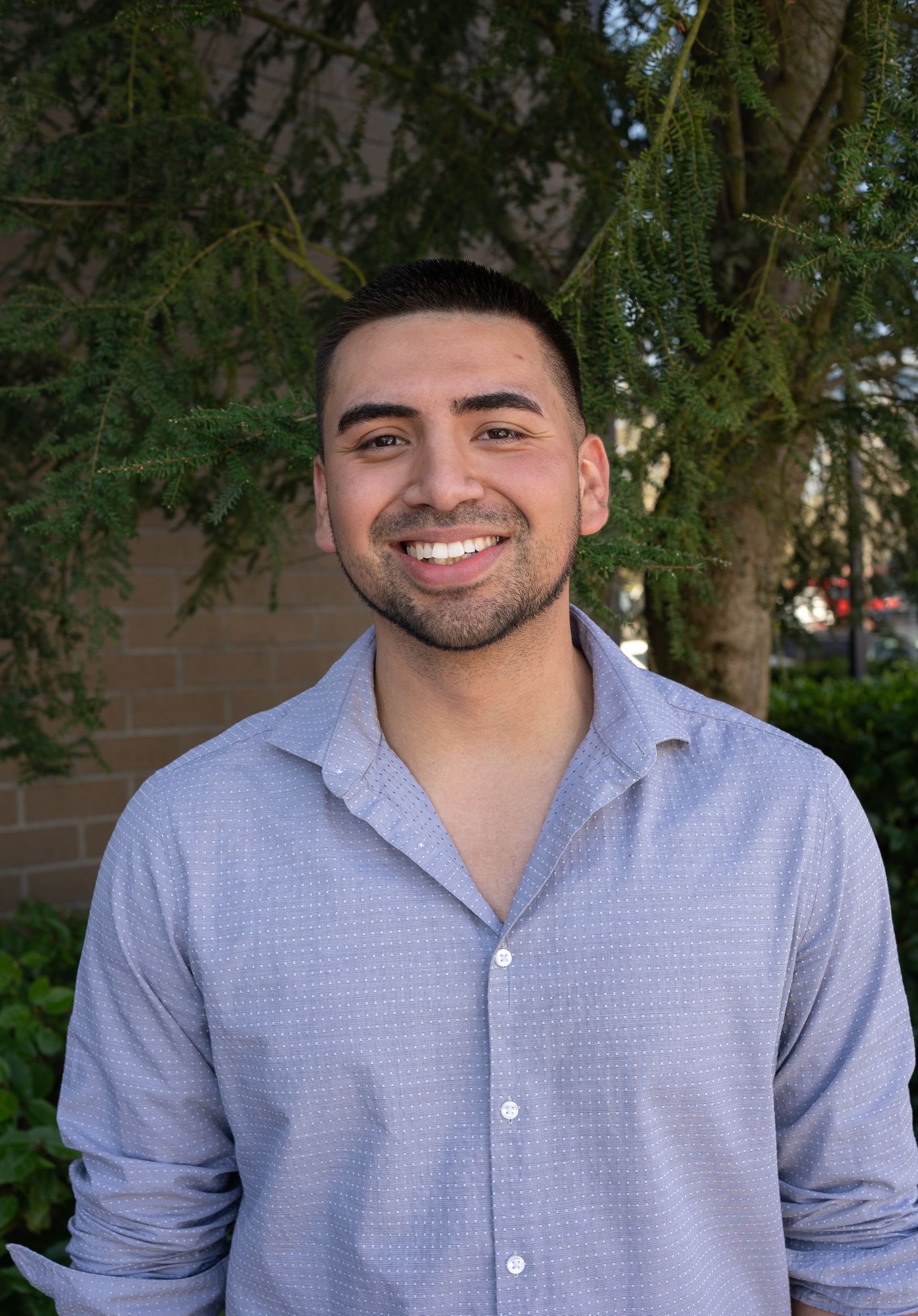 A man in a blue shirt is smiling in front of a tree.