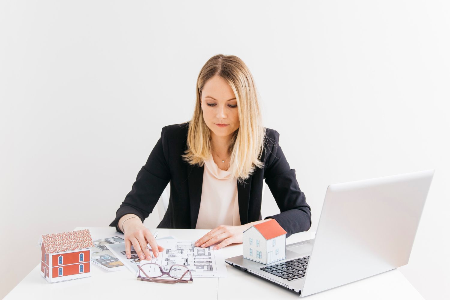 A woman is sitting at a desk with a laptop and a model house.