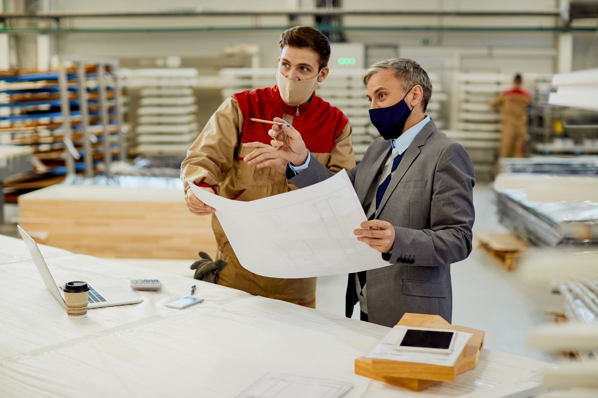 Two men wearing masks are looking at a blueprint in a factory.