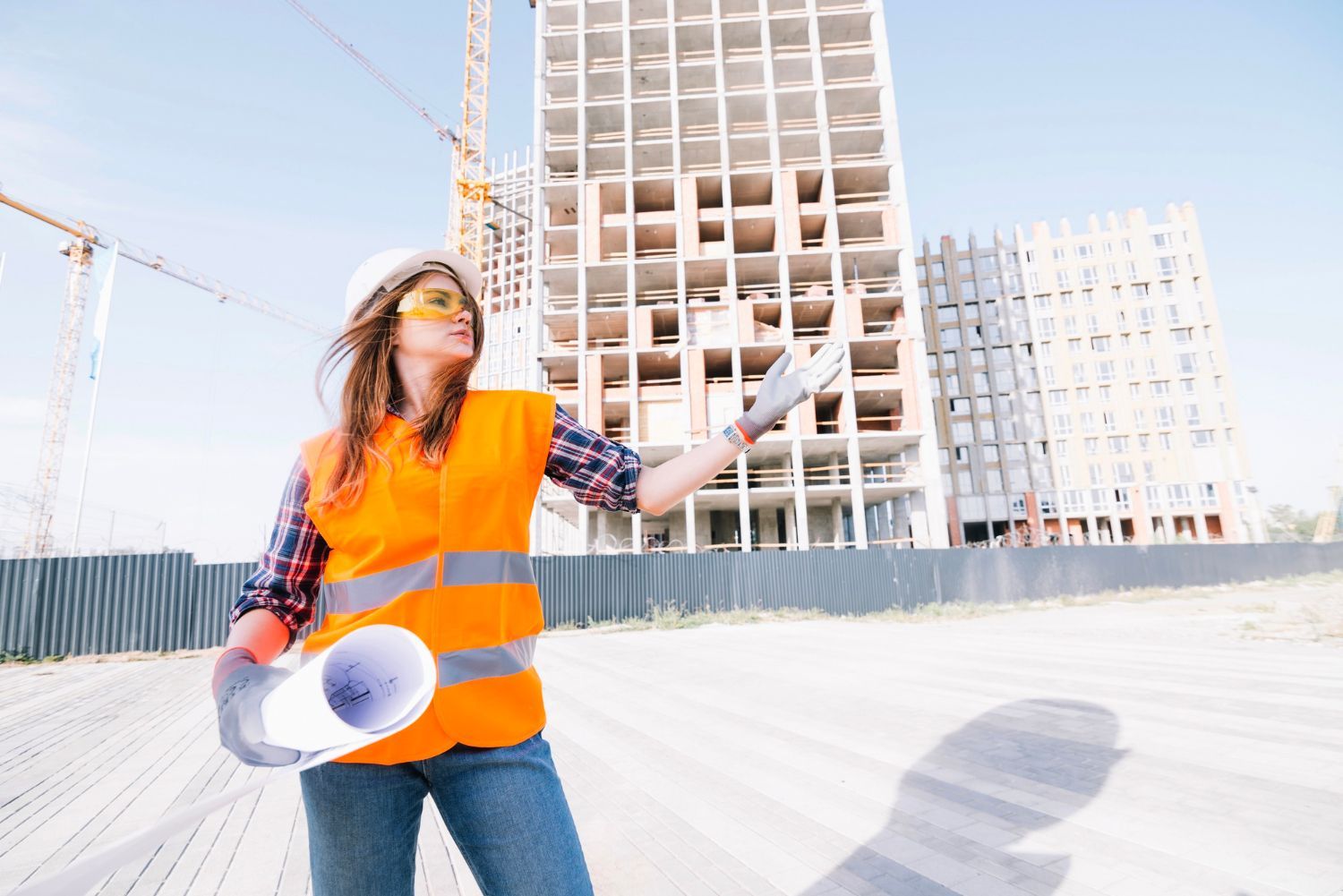 A woman is standing in front of a building under construction holding a blueprint.