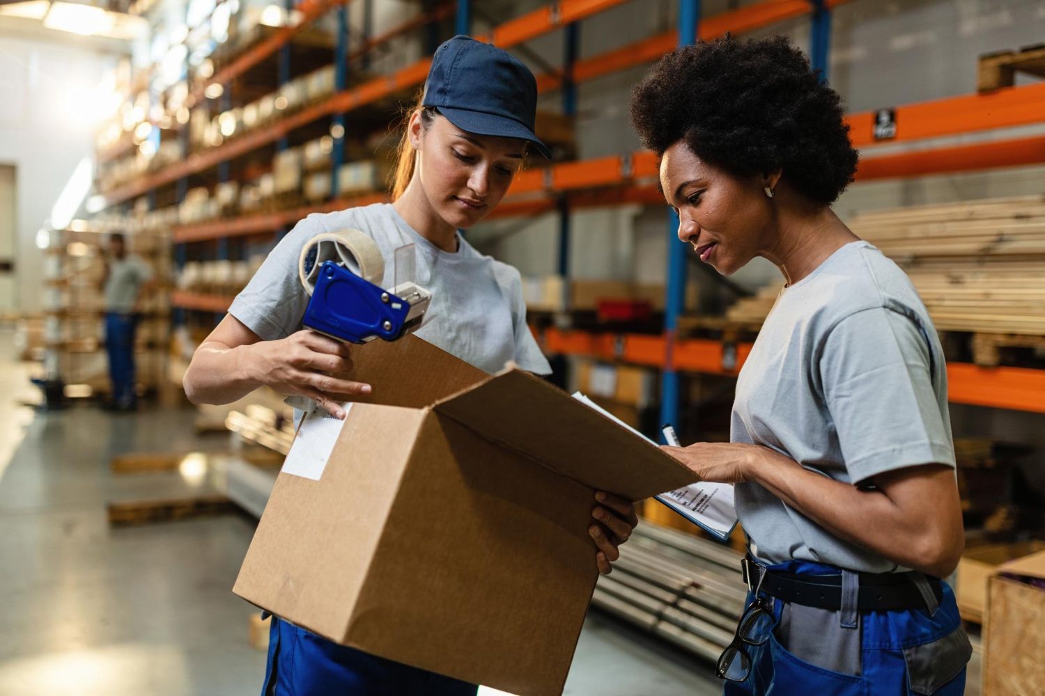 Two women are working in a warehouse , one is holding a box and the other is looking at a clipboard.