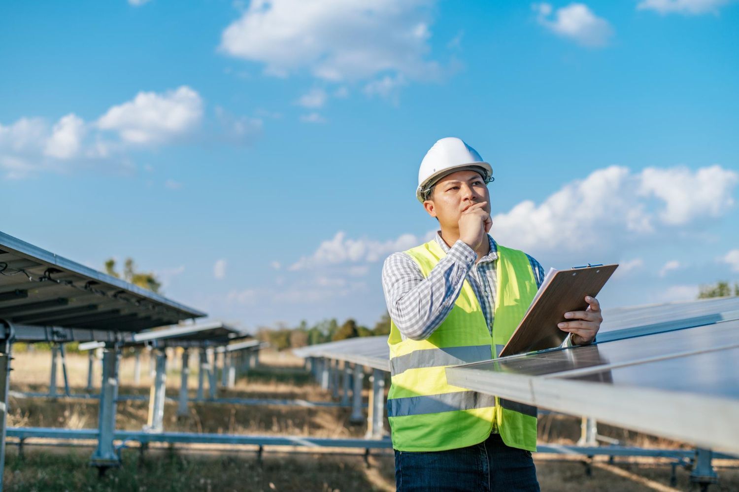 A man in a hard hat is holding a clipboard and looking at a solar panel.