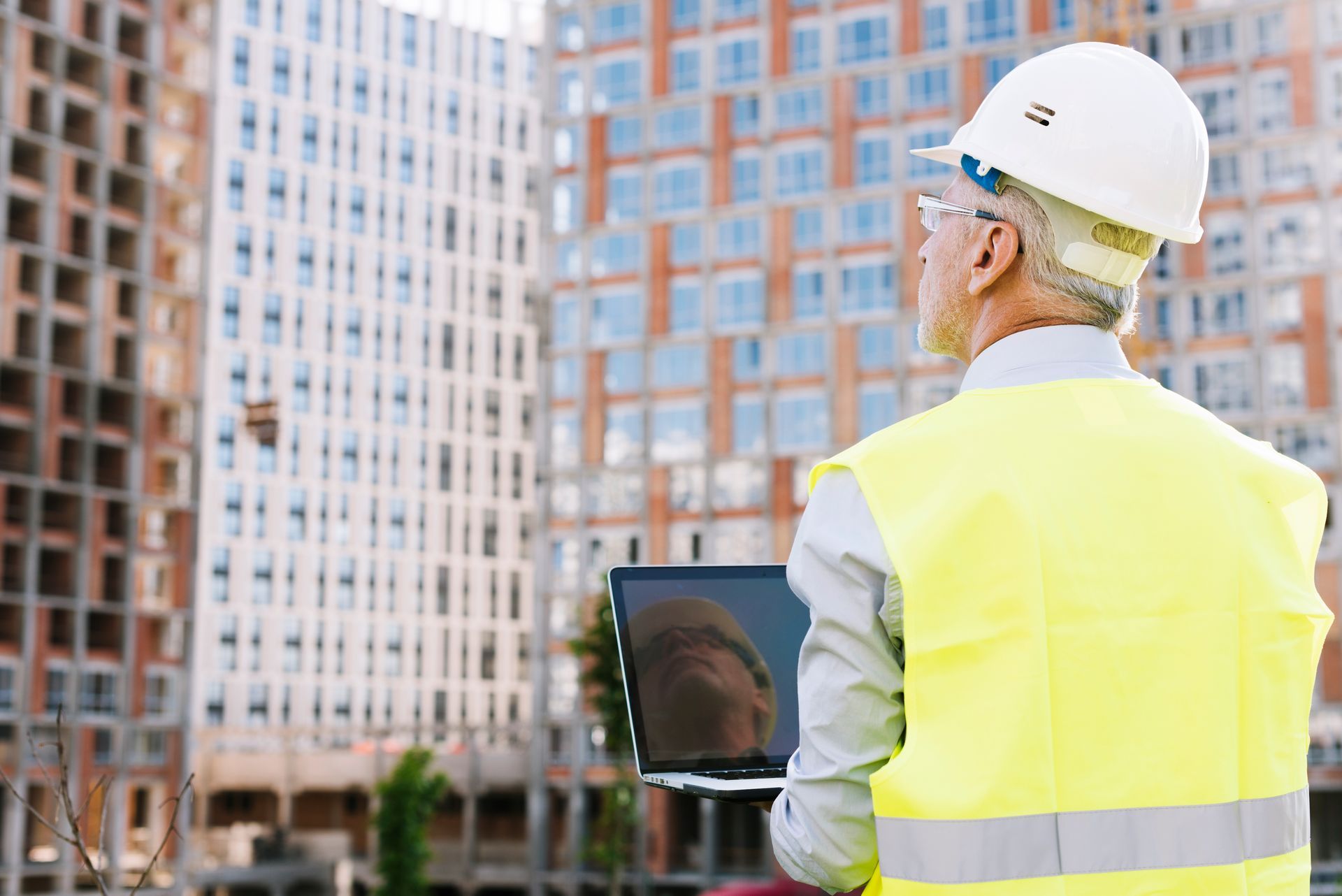 A construction worker is looking at a laptop computer while standing in front of a building.