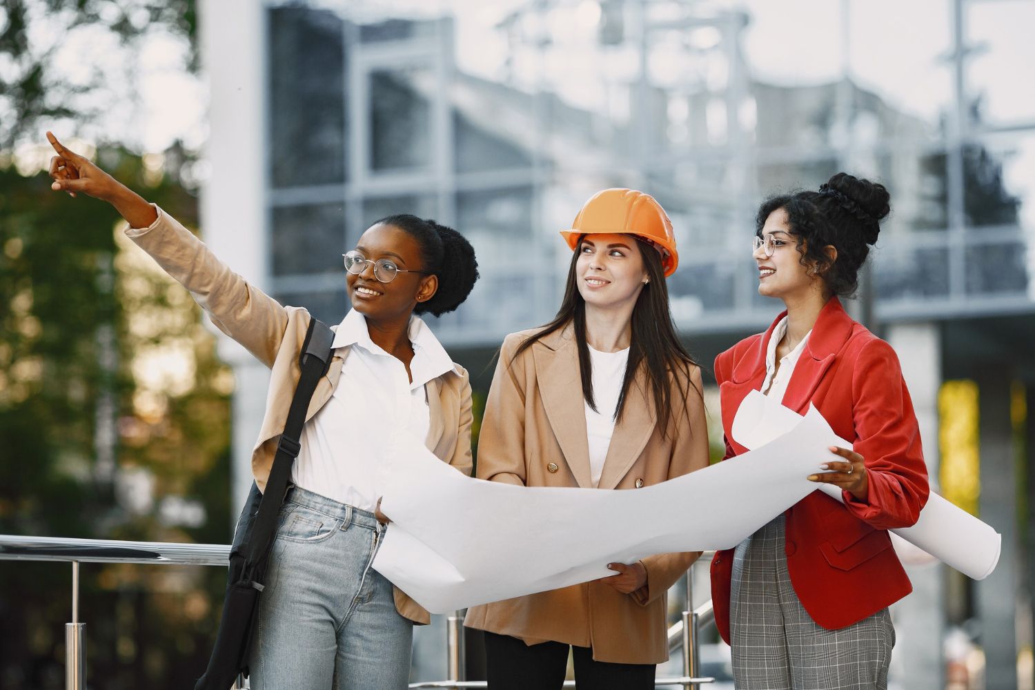 Three women are standing next to each other in front of a building holding blueprints.