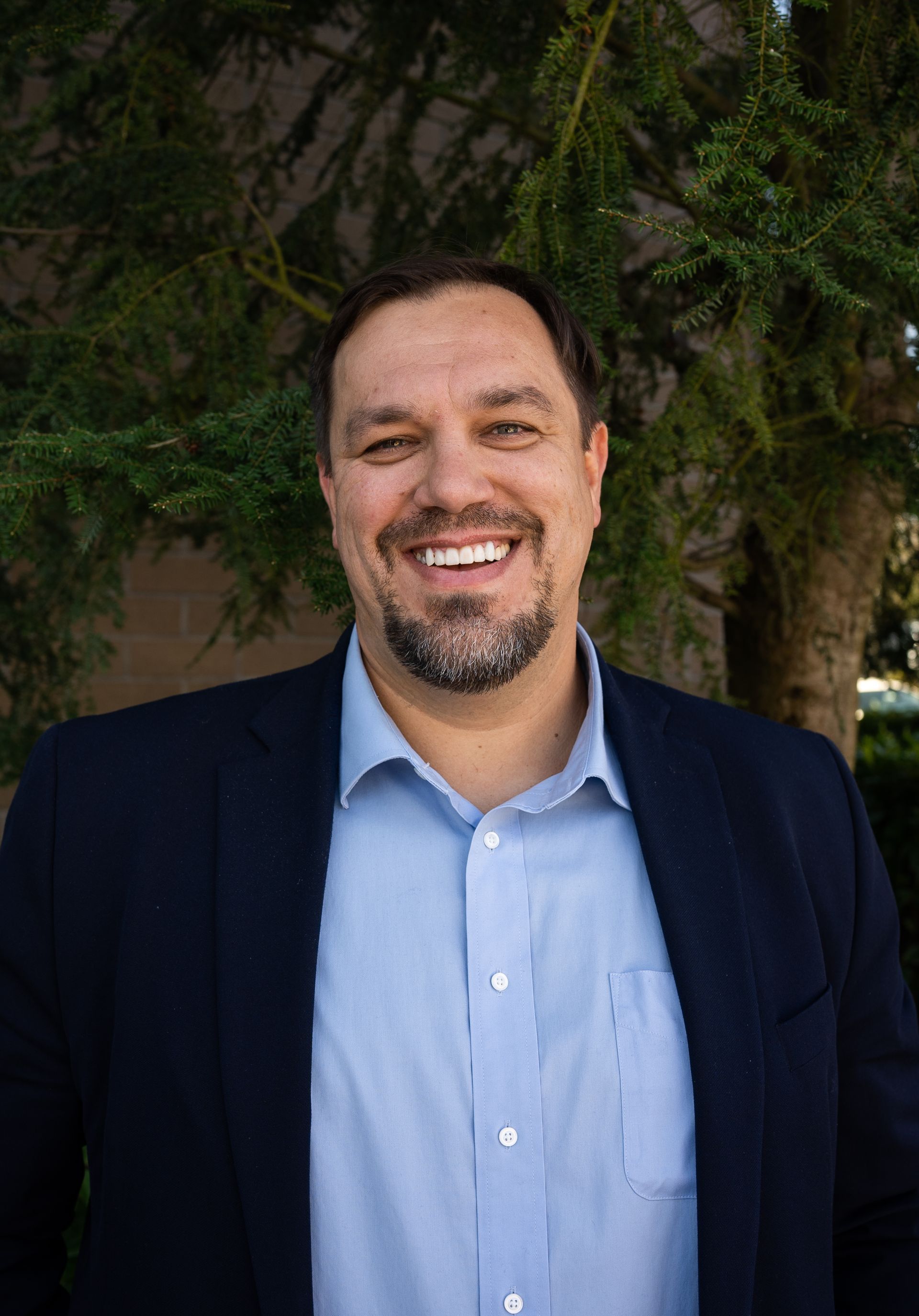 A man in a suit and blue shirt is smiling in front of a tree.