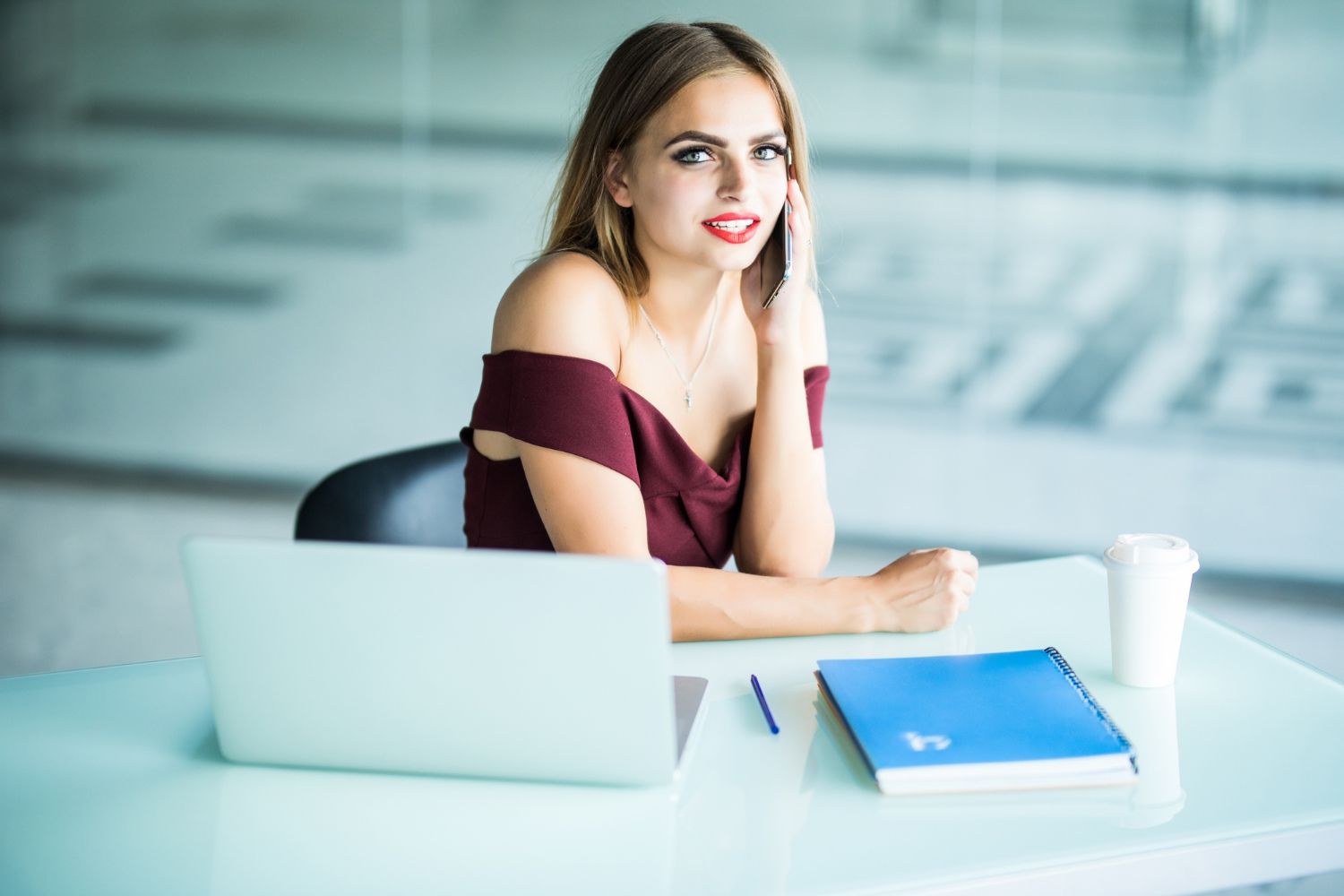 A woman is sitting at a desk with a laptop and talking on a cell phone.