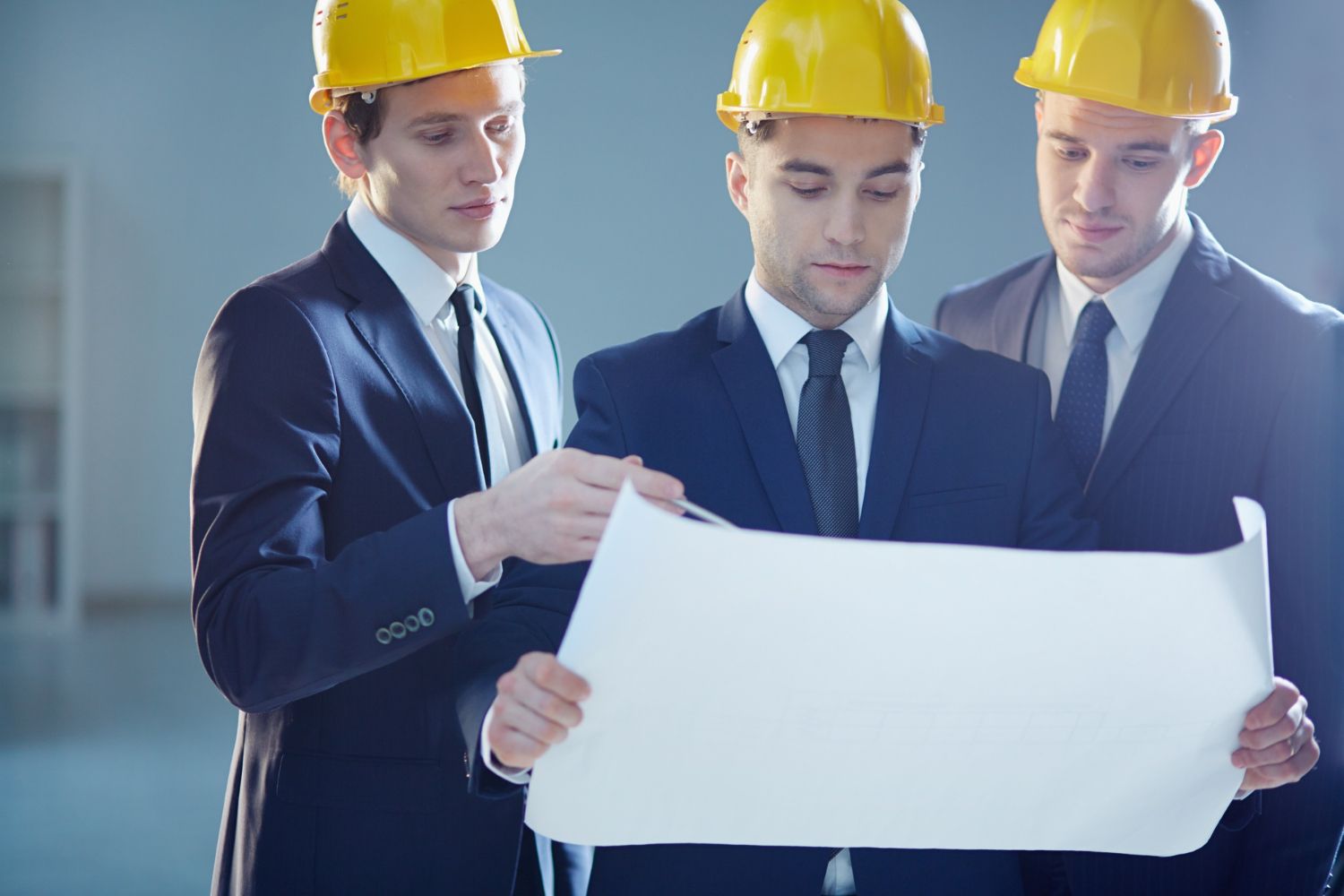 Three men in suits and hard hats are looking at a blueprint.