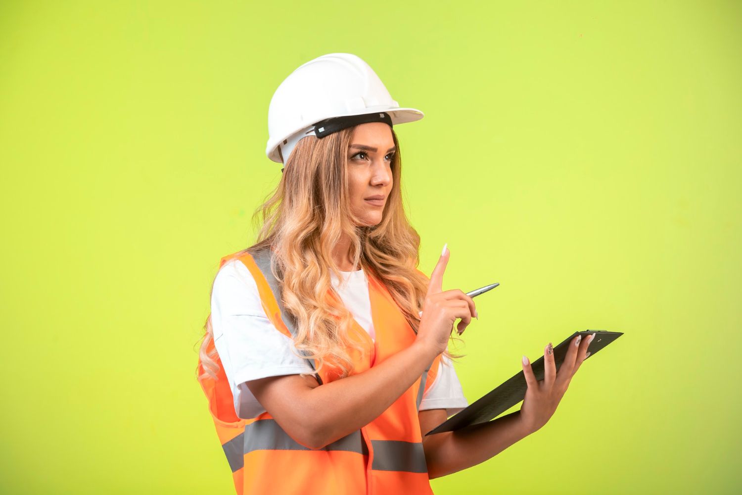 A woman in a hard hat and safety vest is holding a clipboard and pointing at it.