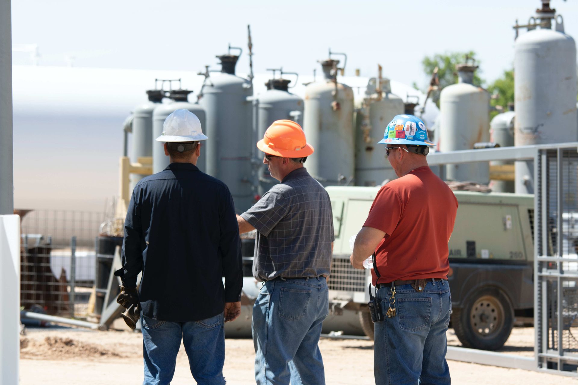 Three men wearing hard hats are standing in front of a factory