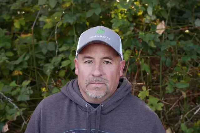 A man wearing a hat and a hoodie is standing in front of a bush.