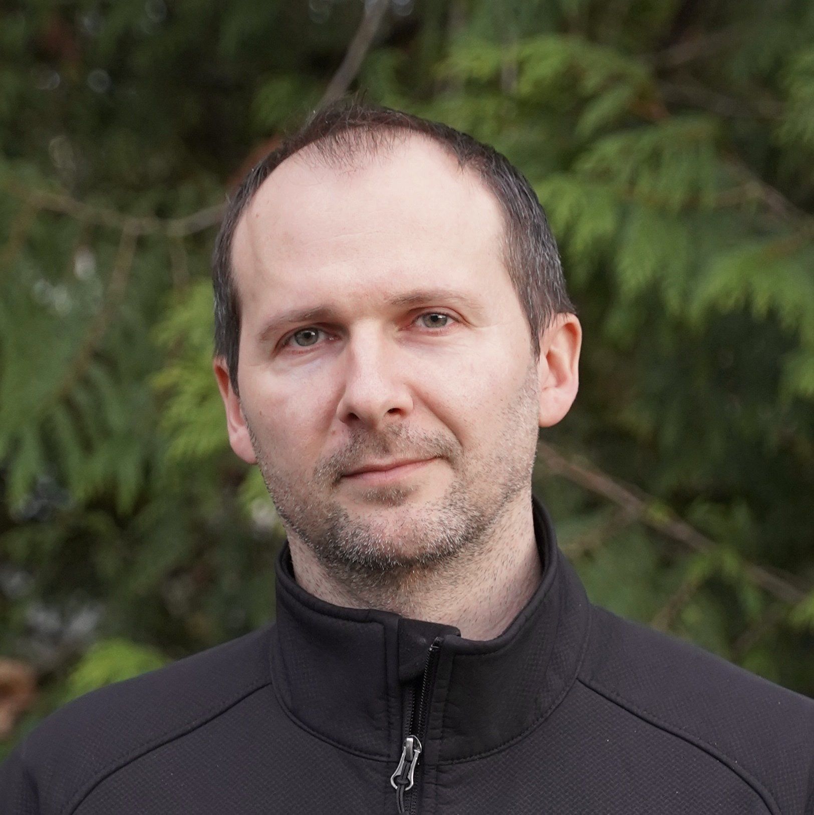 A man with a beard wearing a black jacket is standing in front of a tree.