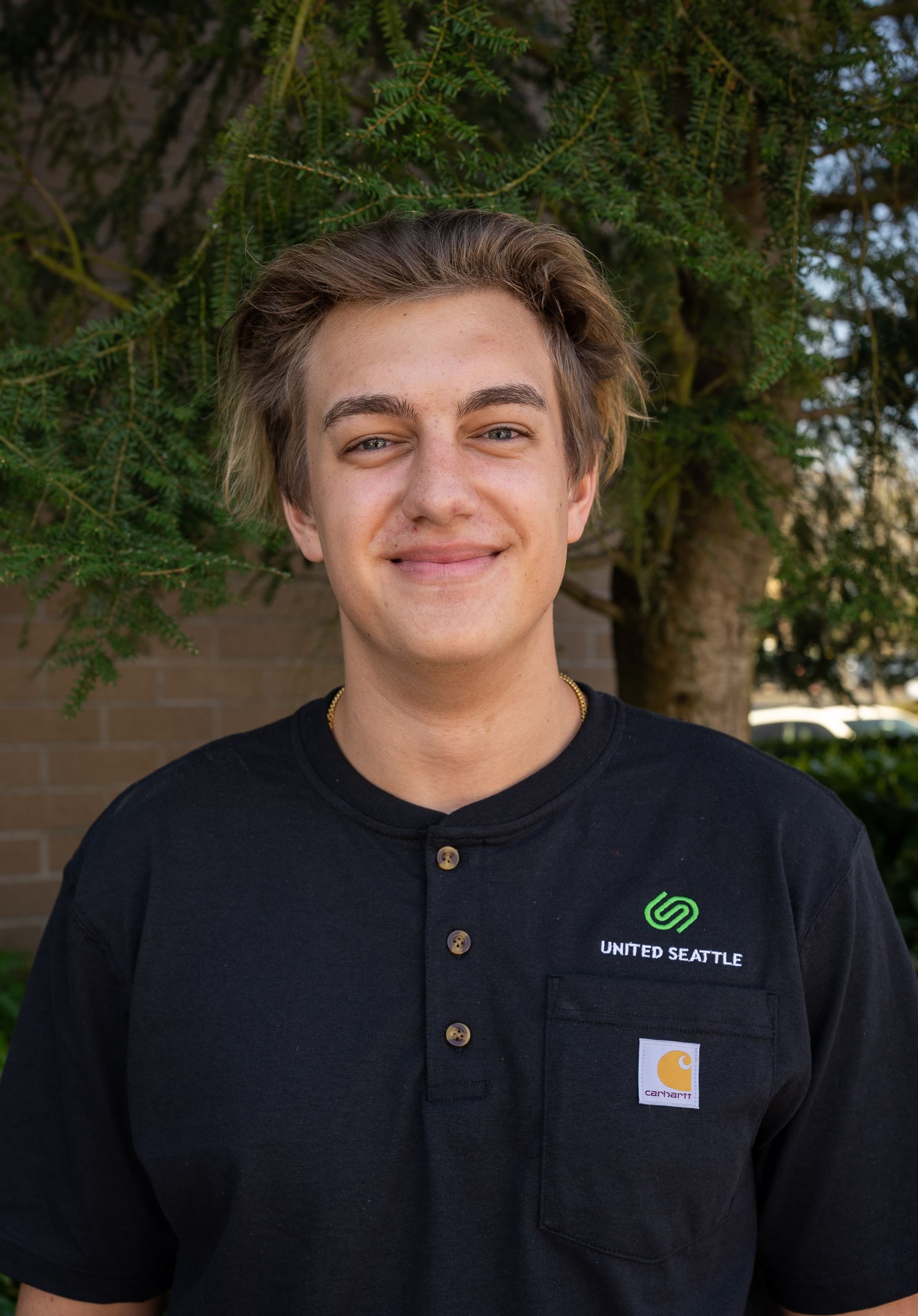 A young man in a black shirt is smiling in front of a tree.