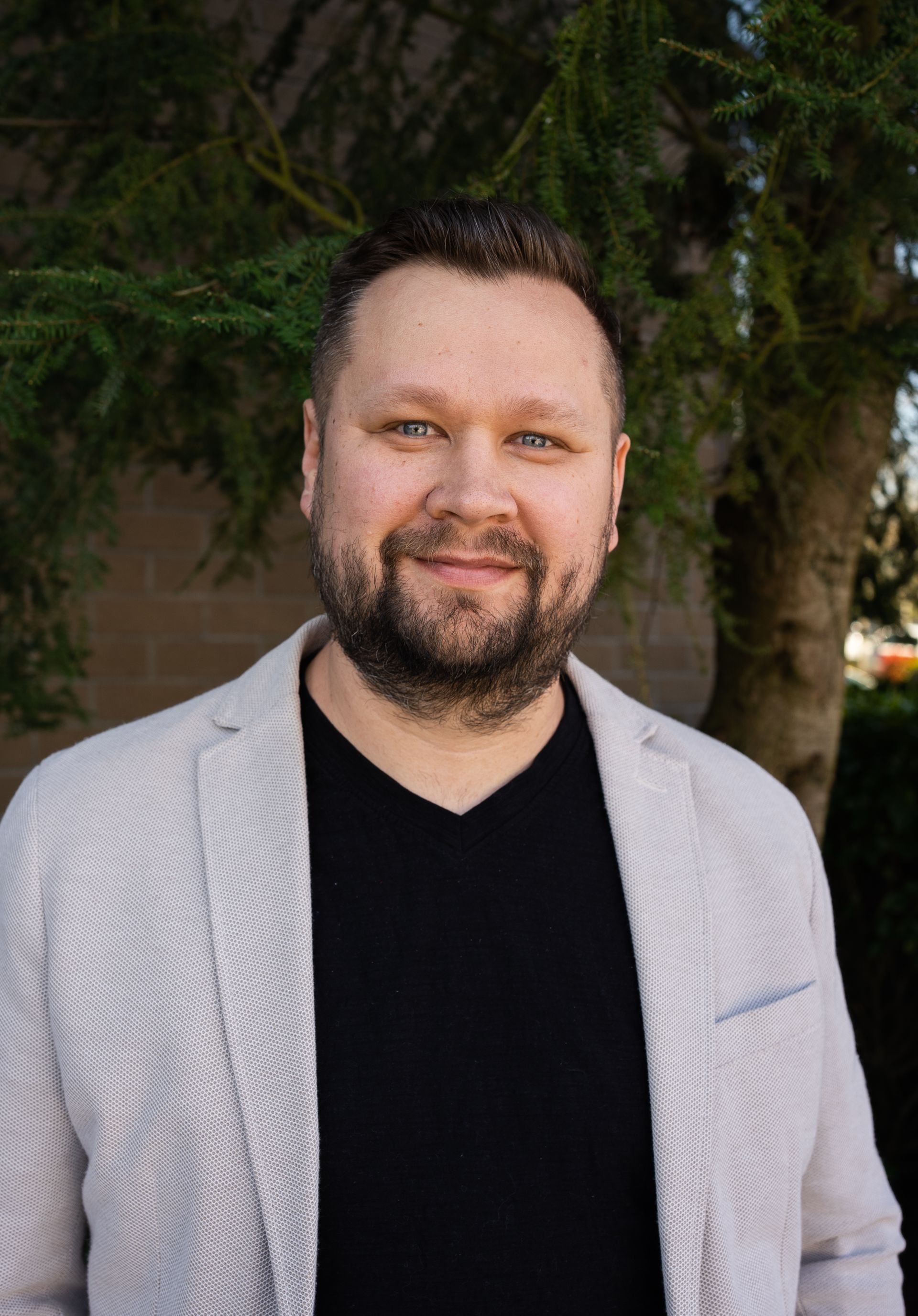 A man with a beard wearing a white jacket and a black shirt is standing in front of a tree.