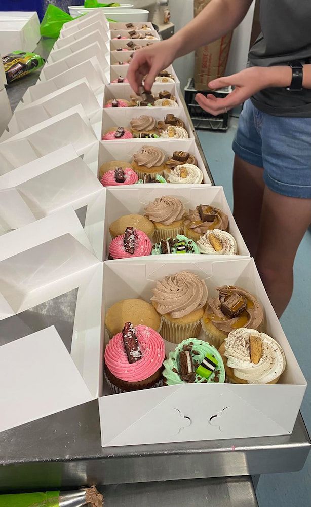 A Woman Is Arranging Cupcakes In Boxes On A Table — Cedar Park Bakehouse in Taranganba, QLD