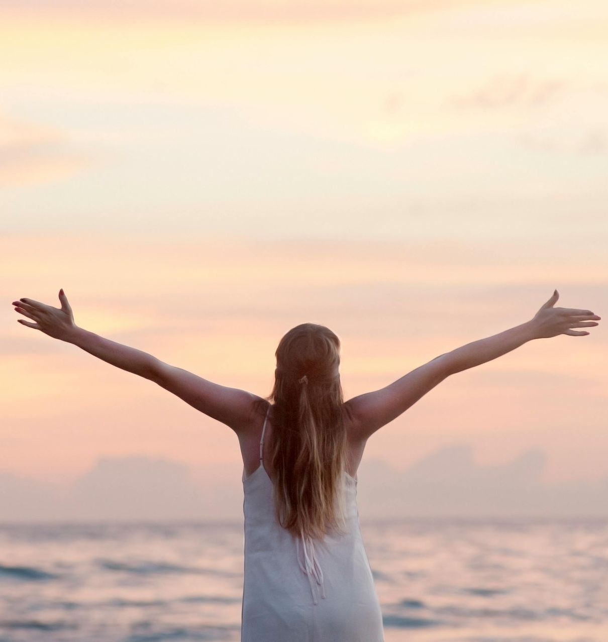 Woman with arms outstretched towards a sunset over the ocean.