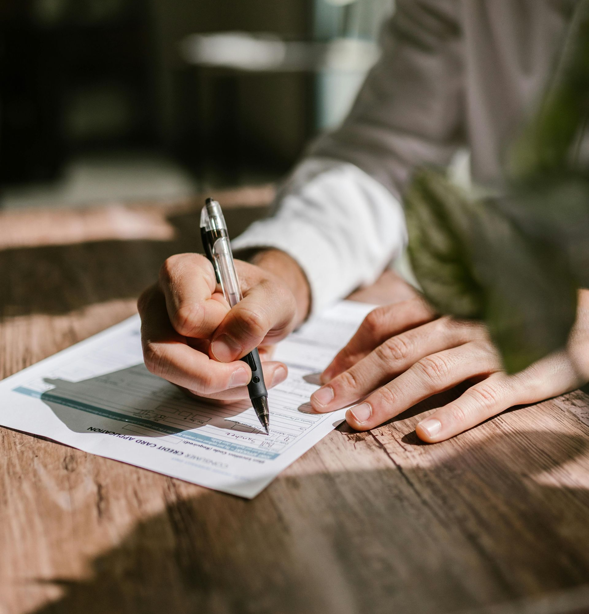 Person writing on a form with a pen at a wooden table in a room with sunlight.