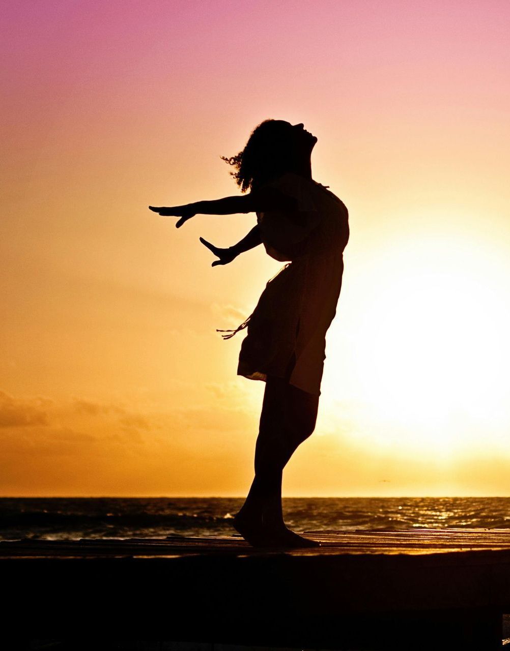 Woman silhouetted against a sunset with arms outstretched, standing on a pier, sun over ocean, embracing freedom.