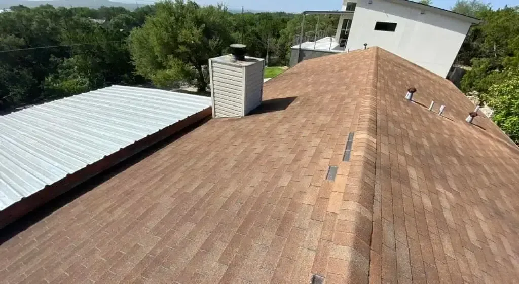 Brown shingle roof with a chimney and metal roof in the background under a bright sky.