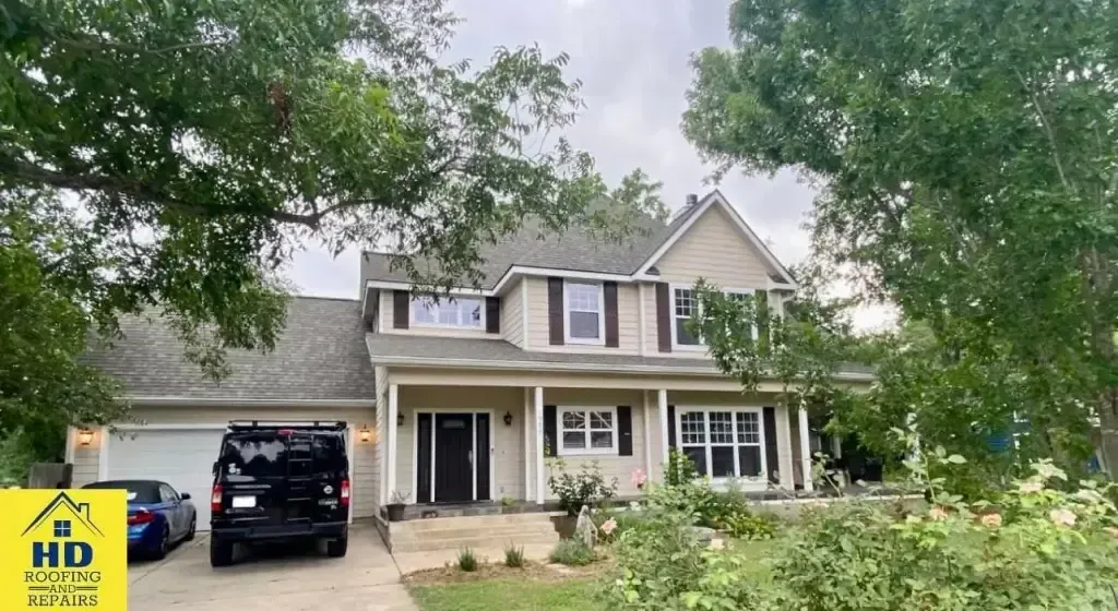 Beige two-story house with a black van and car in the driveway, trees in the yard.