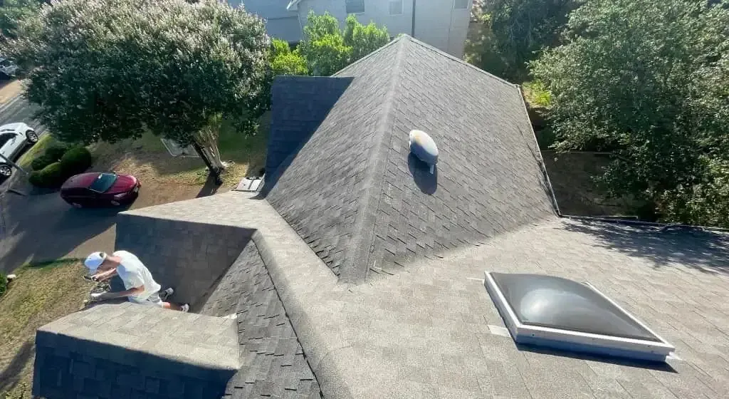 Man working on a dark gray shingled roof with a skylight and vent. Red car and trees in the background.