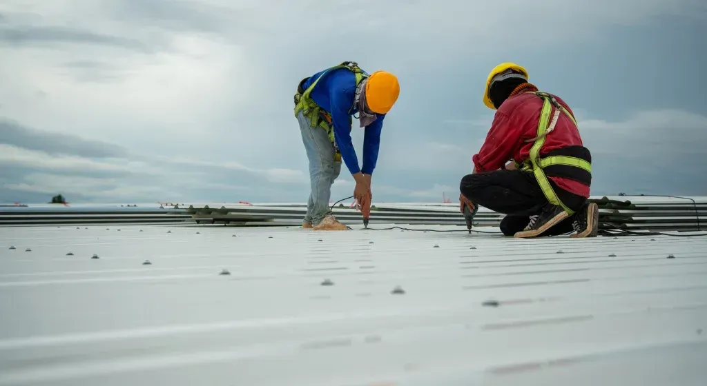 Two construction workers in safety gear working on a white roof under cloudy skies.