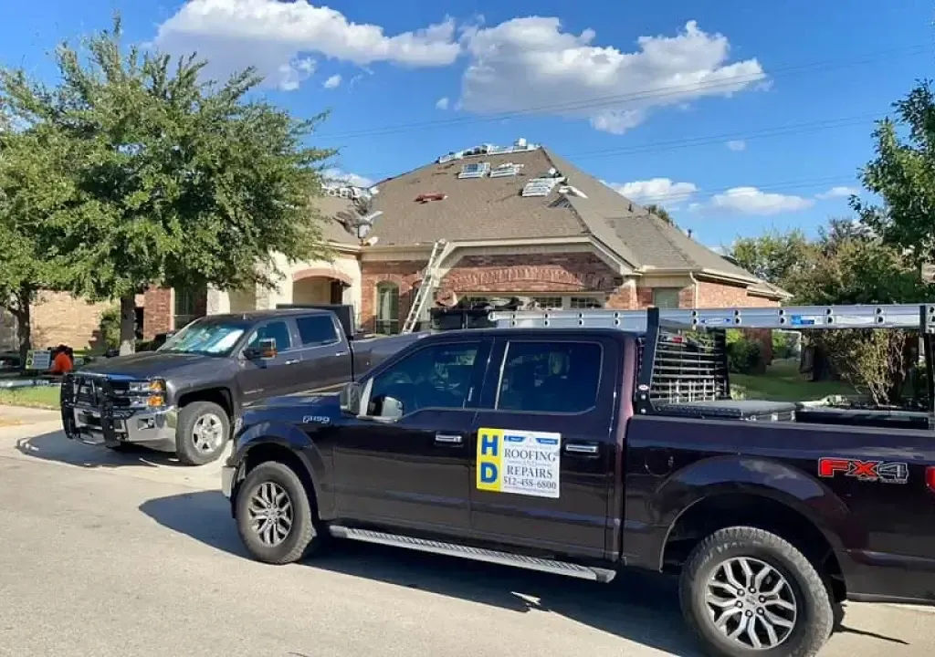 Two pickup trucks parked in front of a house with roof repair in progress. Blue sky with clouds.