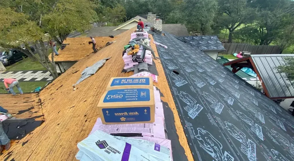 Roofers installing new shingles on a house roof. Boxes of materials and equipment are present.