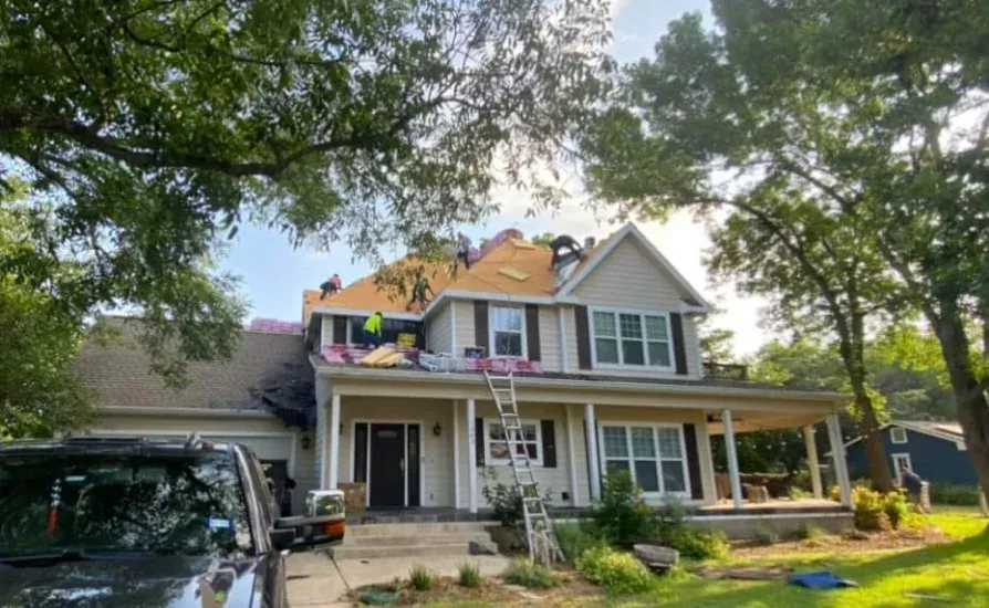 Workers replacing roof shingles on a two-story house with a porch. Trees surround the house, and a truck is parked nearby.